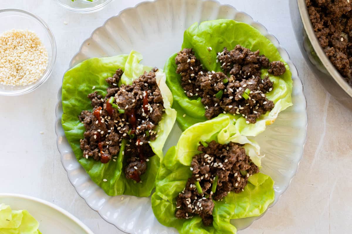 Three lettuce leaves topped with ground meat, chopped green onions, and sesame seeds are arranged on a clear plate, with a small bowl of sesame seeds nearby.