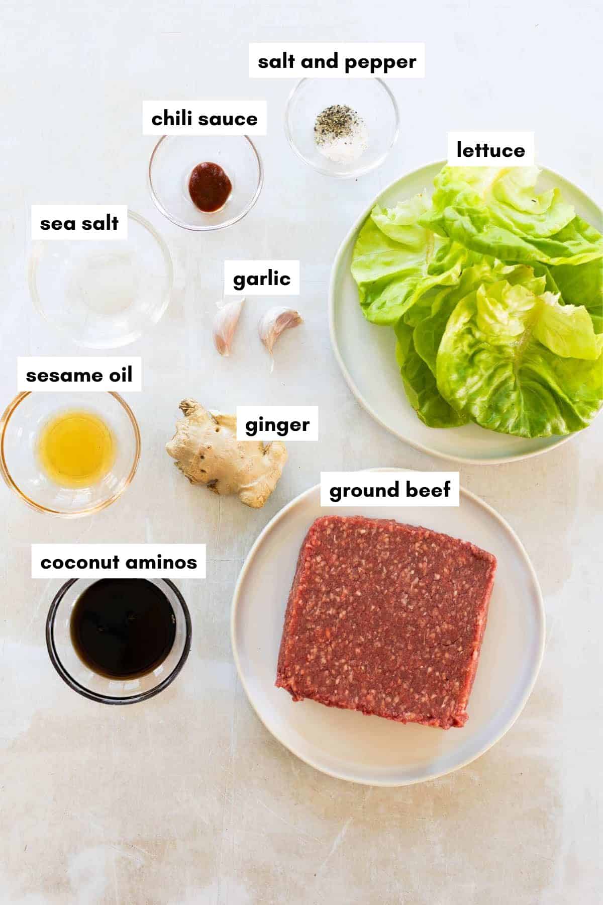 Ingredients ground beef lettuce wraps recipe arranged on a light surface: lettuce, ground beef, ginger, garlic, sea salt, chili sauce, salt and pepper, sesame oil, and coconut aminos in small bowls.