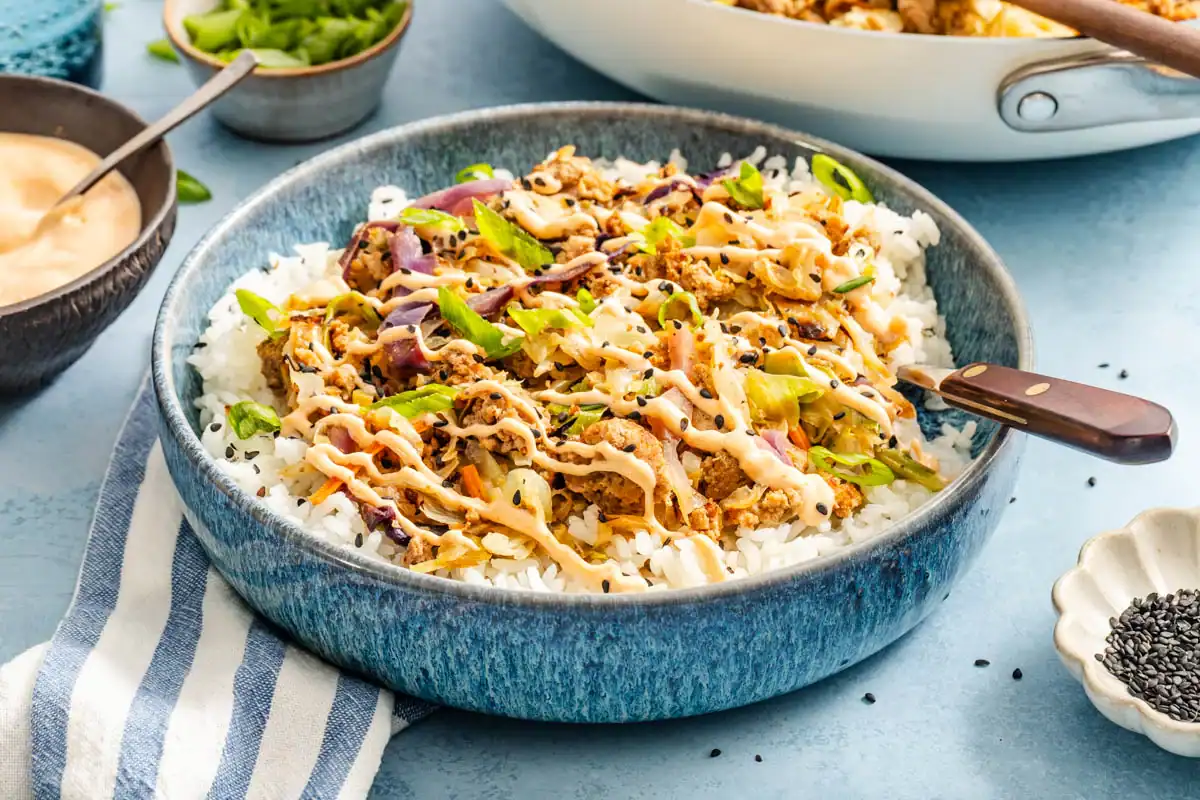 A blue bowl of egg roll in a bowl with ground meat, cabbage, and white rice, drizzled with spicy mayo and topped with green onions and sesame seeds.