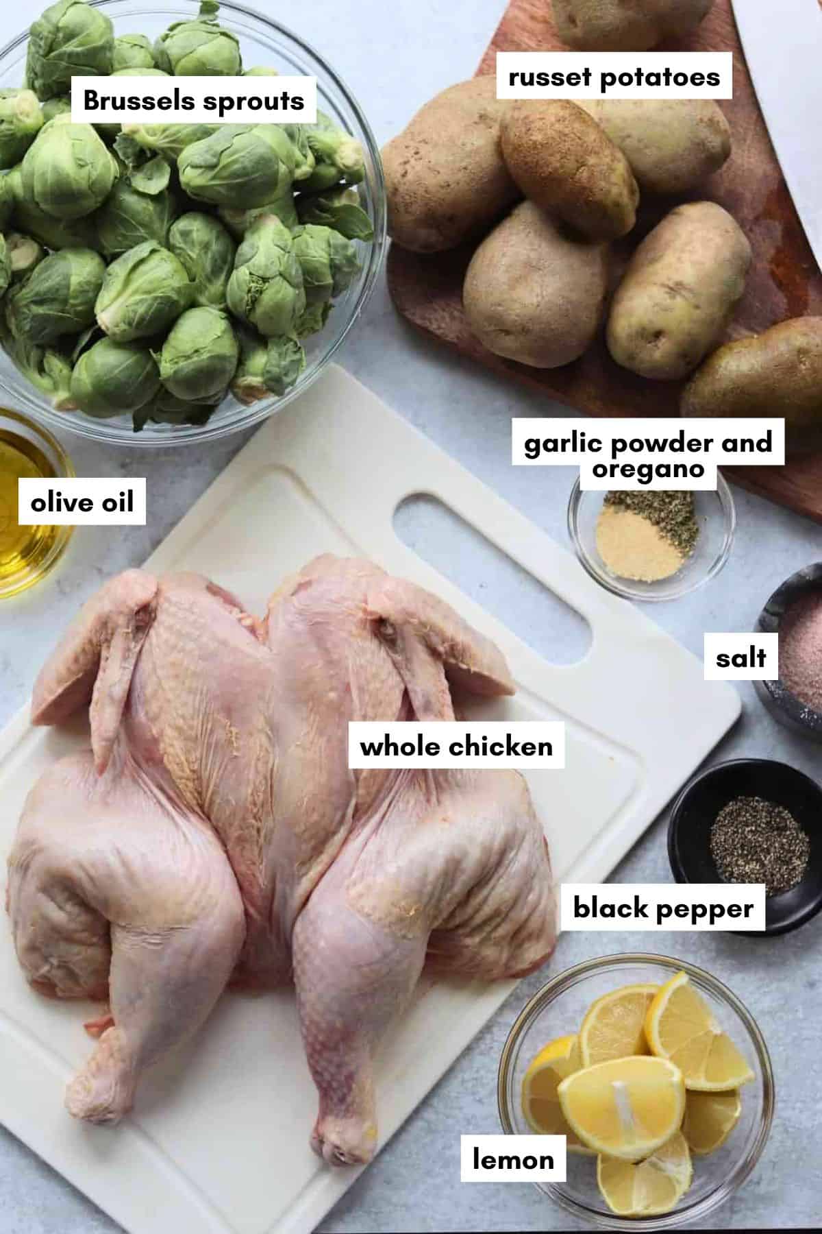 Ingredients for a spatchcock chicken recipe: a whole chicken, Brussels sprouts, russet potatoes, lemon, olive oil, and seasonings including salt, black pepper, garlic powder, and oregano.