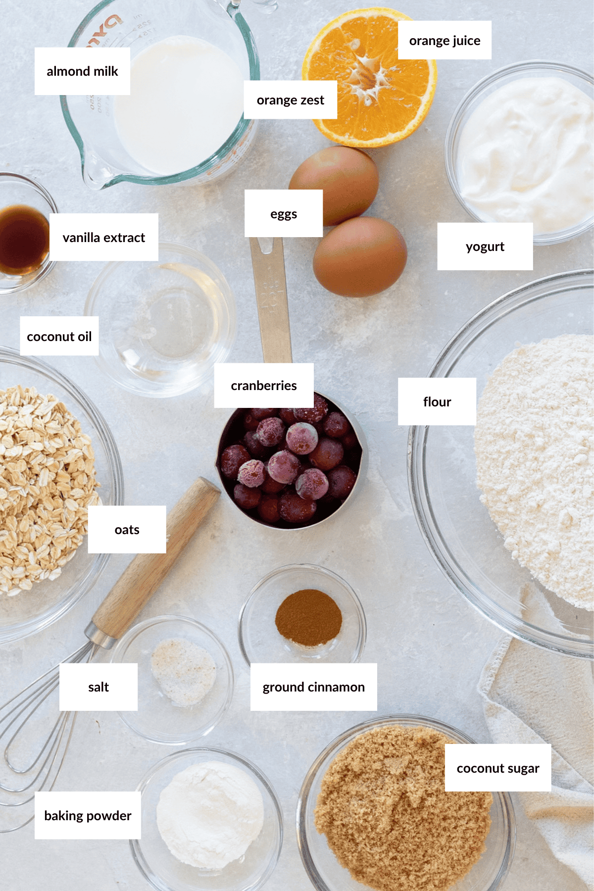 A flat lay of labeled baking ingredients including flour, oats, eggs, cranberries, coconut oil, almond milk, orange juice, zest, yogurt, sugar, baking powder, cinnamon, vanilla, and salt.