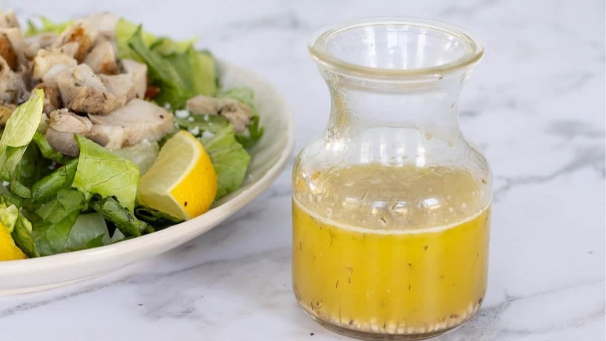 A small glass carafe of lemon vinaigrette salad dressing next to a bowl of salad topped with chicken breast.