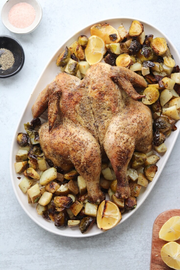 Roast spatchcock chicken on a platter with roasted potatoes, Brussels sprouts, chopped lemons, and seasoning bowls nearby.