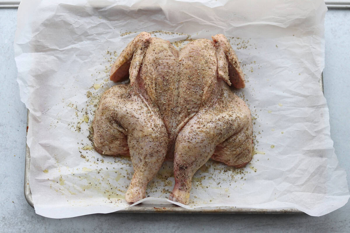 A raw, butterflied chicken seasoned with spices is placed on parchment paper on a baking sheet.