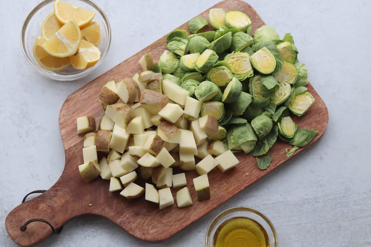 Chopped potatoes and halved Brussels sprouts on a wooden board, with a bowl of lemon wedges and a small bowl of olive oil nearby.