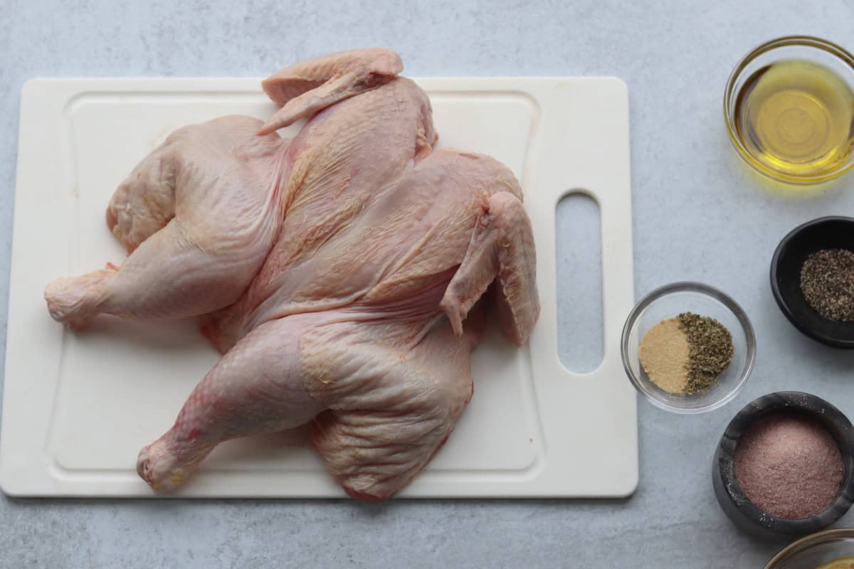 Raw spatchcocked chicken on a white cutting board with bowls of oil, salt, pepper, and seasonings on a light surface.