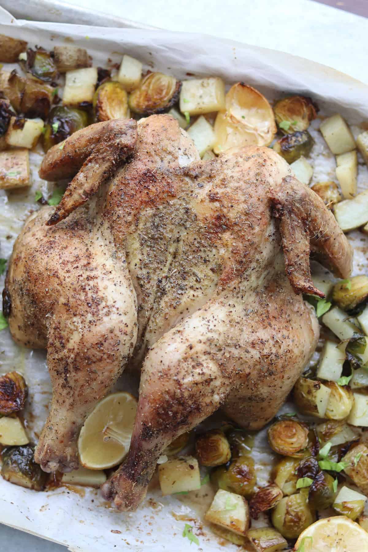 Roasted spatchcock chicken seasoned with herbs, surrounded by roasted Brussels sprouts, potatoes, and lemon wedges on a parchment-lined baking tray.