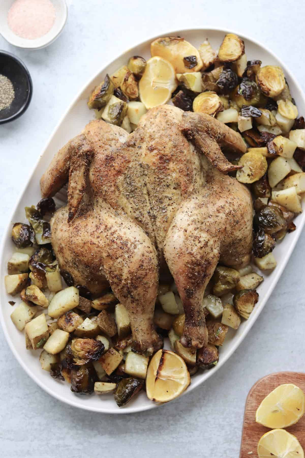 A seasoned roasted spatchcock chicken is served on a platter with roasted potatoes, Brussels sprouts, and lemon wedges. Bowls of salt and pepper are nearby.