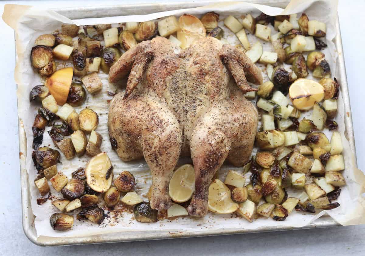 A roasted spatchcocked chicken on a baking sheet, surrounded by roasted potatoes, Brussels sprouts, and lemon wedges.