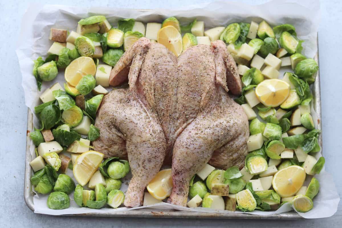 A raw spatchcocked chicken seasoned with spices is surrounded by chopped Brussels sprouts, potatoes, and lemon slices on a parchment-lined baking sheet.