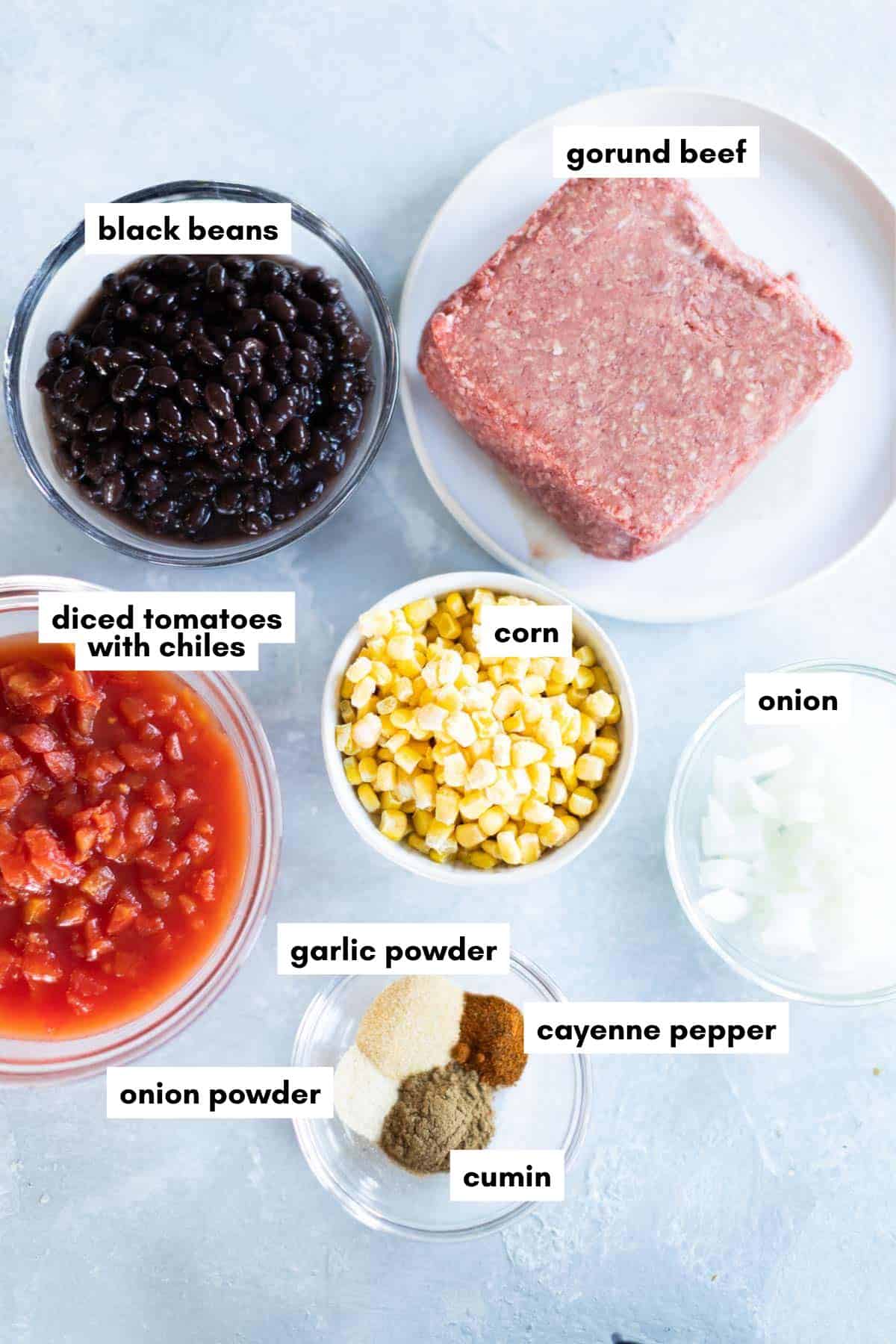 Ground beef, black beans, diced tomatoes with chiles, corn, chopped onion, and small bowls of garlic powder, onion powder, cumin, and cayenne pepper are arranged on a light surface—perfect ingredients for a hearty ground beef taco soup.